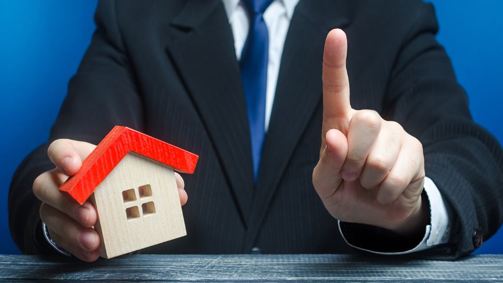 スーツを着た男性が左手で正面に向かって指差し、右手に家の模型を持っている